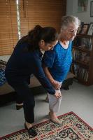 Older female doing a lunge with support from a ffemale physio in the loungeroom
