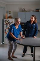 Older female doing exercises holding a side bench in the kitchen supported by a female physio