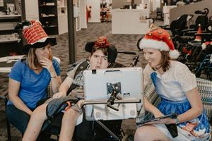 Two females sitting either side of a male in a wheelchair, all wearing Christmas hats & looking at a mounted ipad