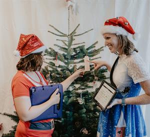 Two females in Christmas garb holding AAC devices standing in front of a Christmas tree pointing and holding a tag