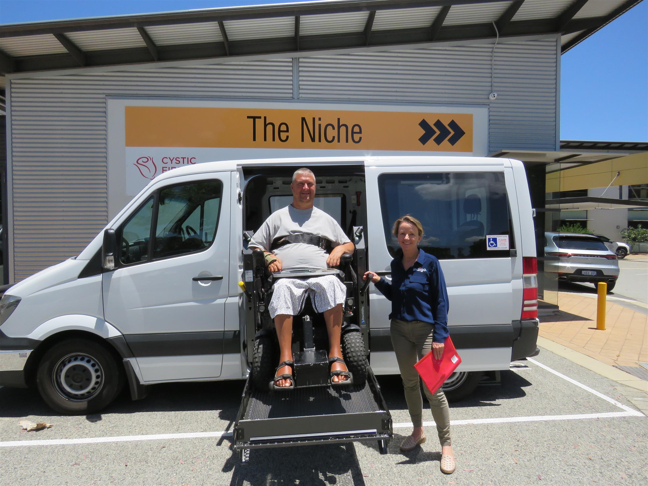 Male in wheelchair on car hoist with female Indigo staff  standing next to hoist