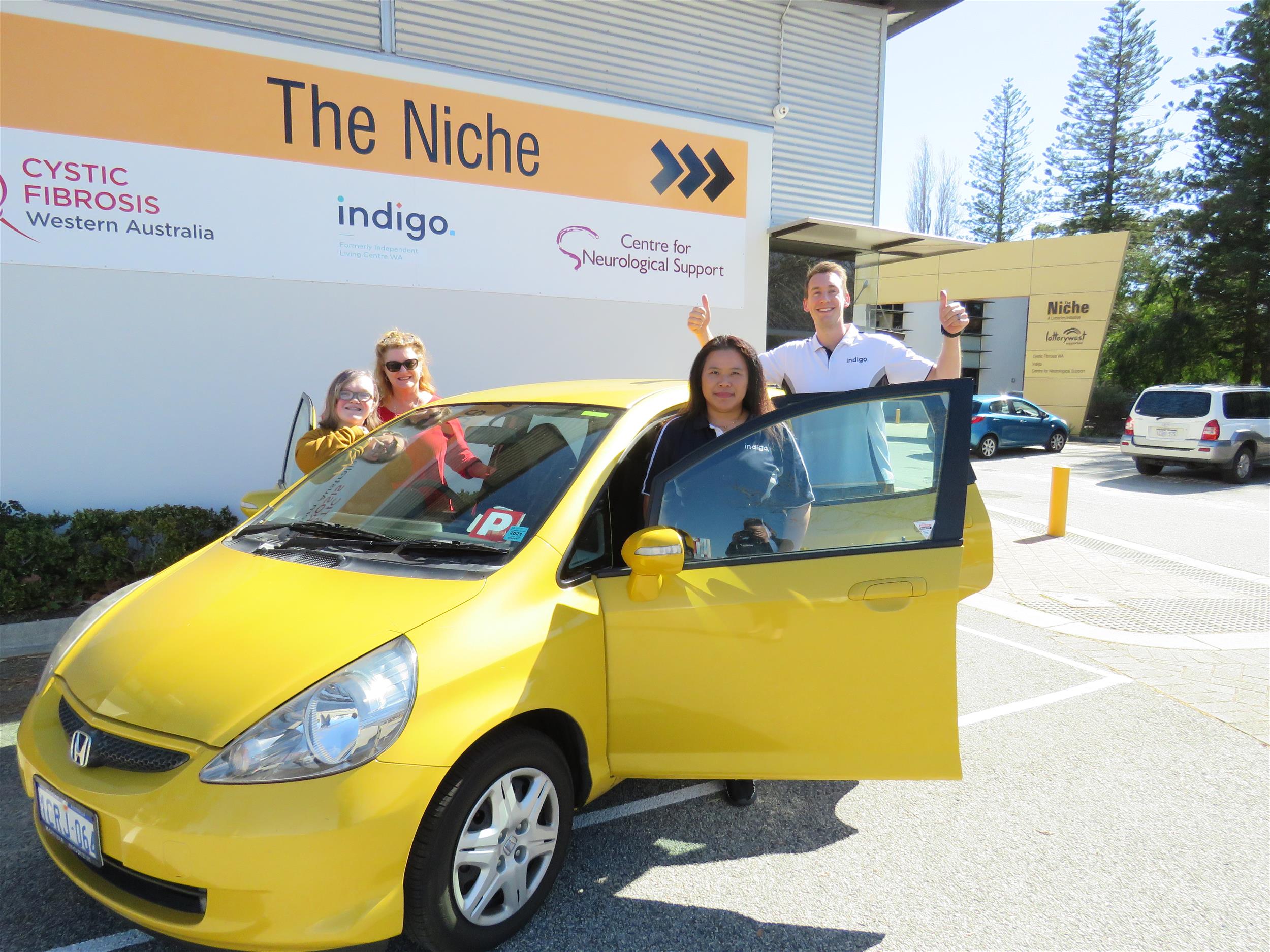 Indigo staff and two female customers all standing in the doors of a yellow car at the front of the Niche