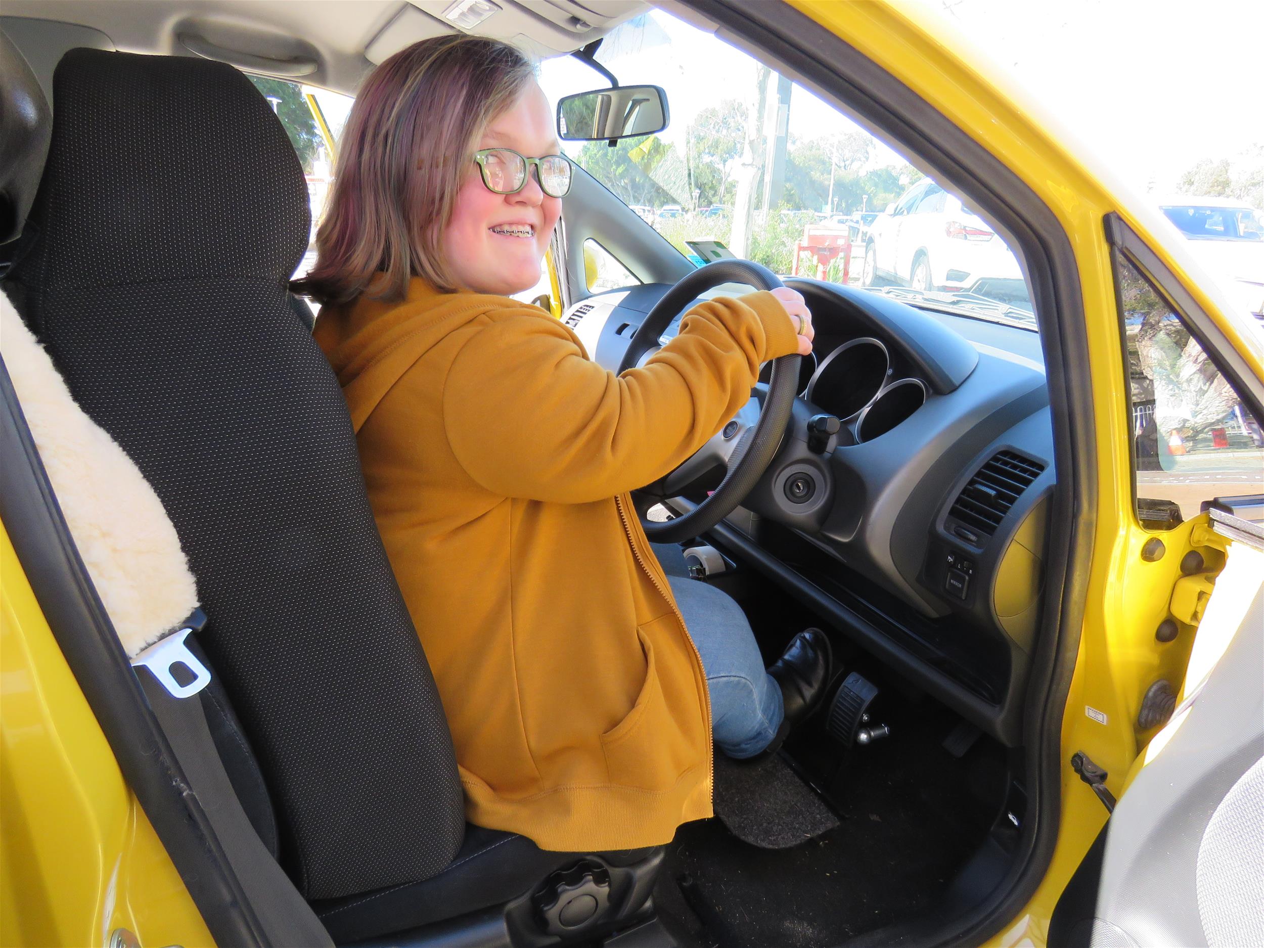 Young adult female in yellow cardigan sitting in driver's seat with hands on the wheel and car door open