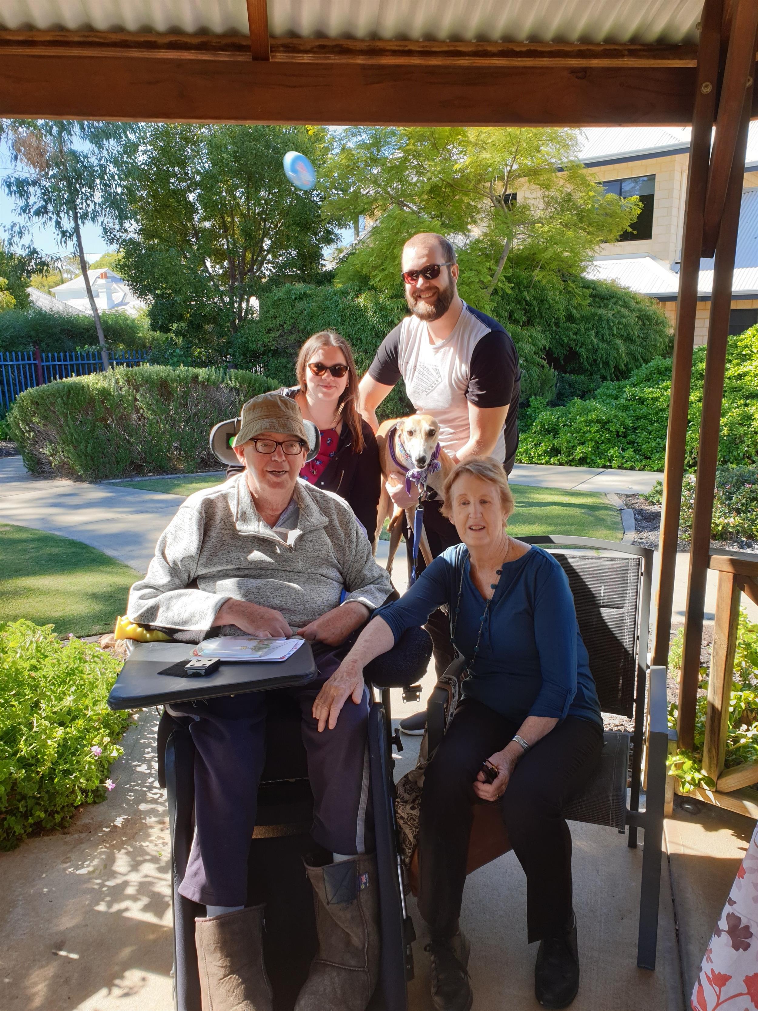 Family of four, 2 males (one in a wheelchair) and 2 females (one seated in a chair next to male in wheelchair) posing outside with their pet greyhound