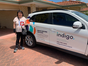Female in black pants and white t-shirt holding P plates and driver's licence standing next to Indigo's Driving team's vehicle