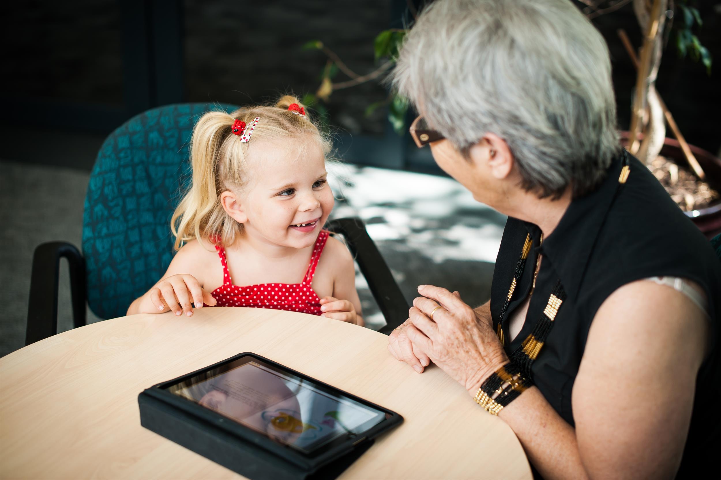 smiling young girl in red dress looking at an older lady in side profile wearing glasses with a tablet on the table