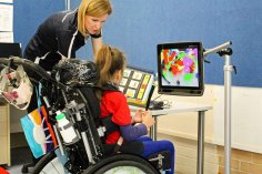 young female in red shit in a wheelchair with headrest with female OT tot he side looking at a mounted computer screen