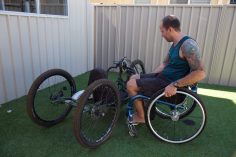 male in t-shirt and shorts in a wheelchair looking at a adjusted racing machine in a backyard