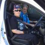 female in sunglasses cap in driver's seat of car with door open and male in the passenger seat looking at camera