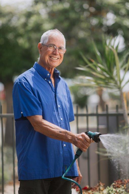 Older male in blue shirt & glasses smiling watering garden