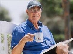 Older male in blue shirt seated on outdoor bench holding cup and a book