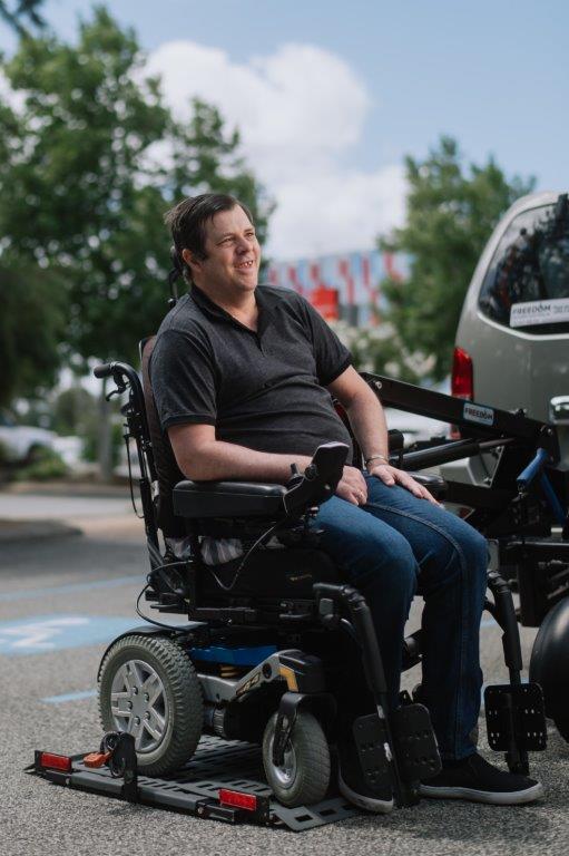 Male in black shirt and jeans in powered wheelchair positioned on lift behind parked vehicle