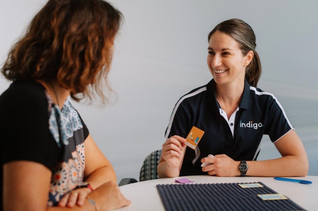female sitting at table wearing indigo shirt holding and showing word card to female in profile sitting at table