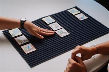  3 rows of picture cards on mat on desk with hand indicating middle row and others hands together on desk