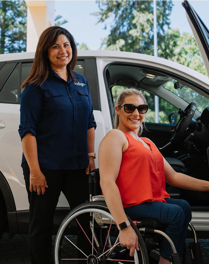 Smiling dark haired OT in Indigo shirt, with blonde, young, fit, NDIS participant next to car