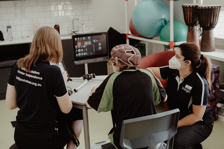 Two female Indigo allied health professionals assisting a student in a powered wheelchair  to use an eyegaze device