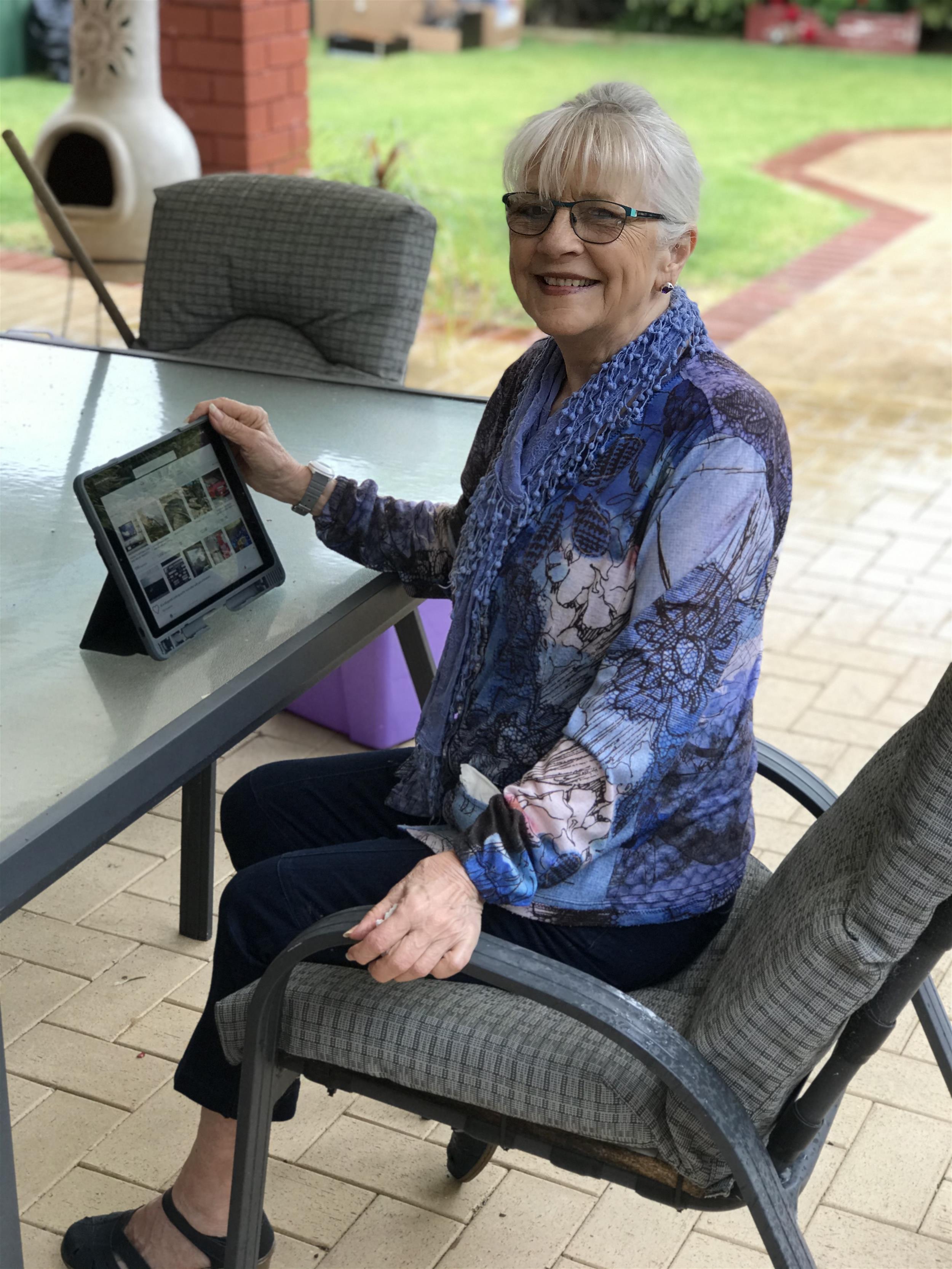 Smartly dressed older lady, with white bun and glasses, smiling. Seated at outdoor table with iPad in front of her.