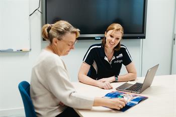 Female Indigo speech pathologists with older female at a desk looking at laptop and brochures
